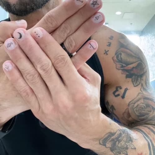 Tattooed hand with short nude press-on nails and minimalist black symbol art โ moon, dots, arrow