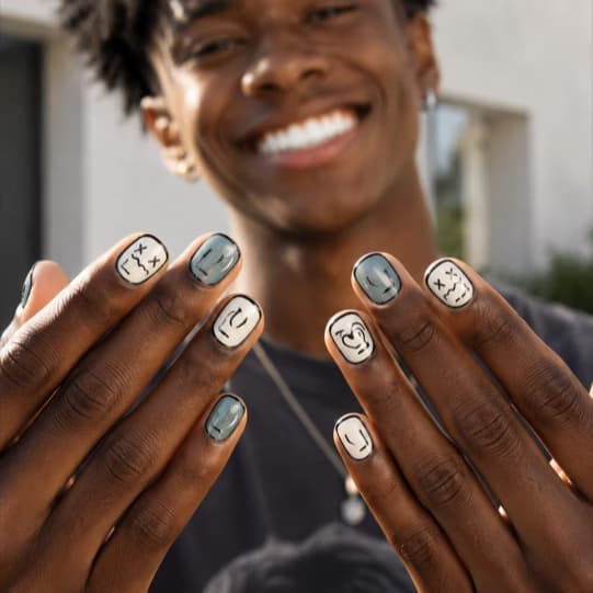 Deep skin-tone hands showing cream and grey press-on nails with hand-drawn doodle faces